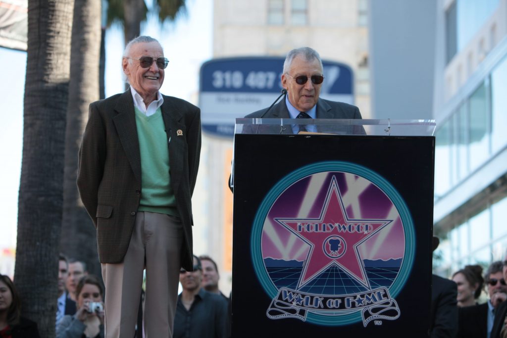 Stan Lee laughing while Gill Champion, standing behind a podium, gives a speech.