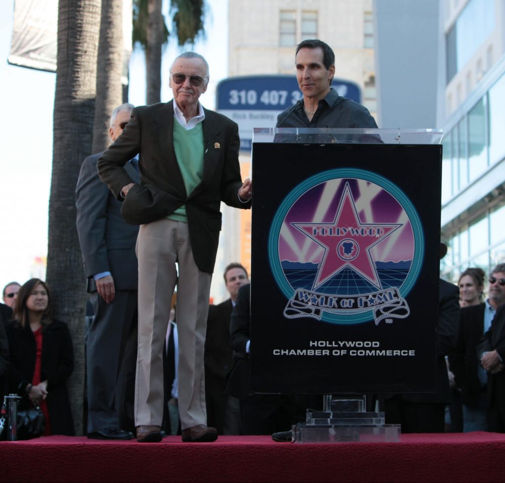 Todd McFarlane giving a speech behind a podium as Stan Lee listens next to him.