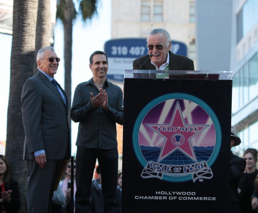 Stan Lee speaking at a podium at this Walk of Fame ceremony with Gill Champion and Todd McFarlane behind him.