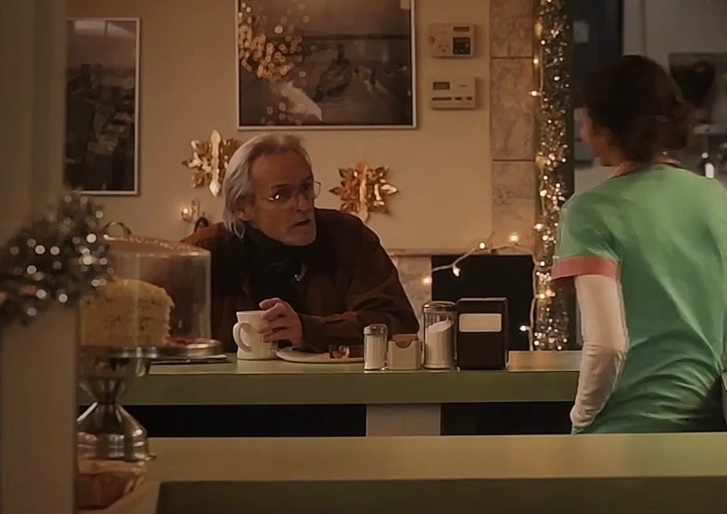 An older man with shaggy white hair sits at a counter holding a white coffee cup