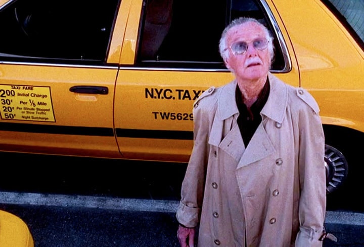 Stan Lee in a beige trenchcoat standing in front of a yellow New York City taxi cab