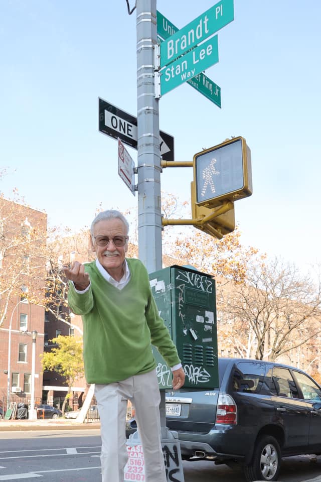 A Stan Lee wax figure in a great sweater and khaki pants with his hand extended underneath a Stan Lee Way street sign on a New York City street.