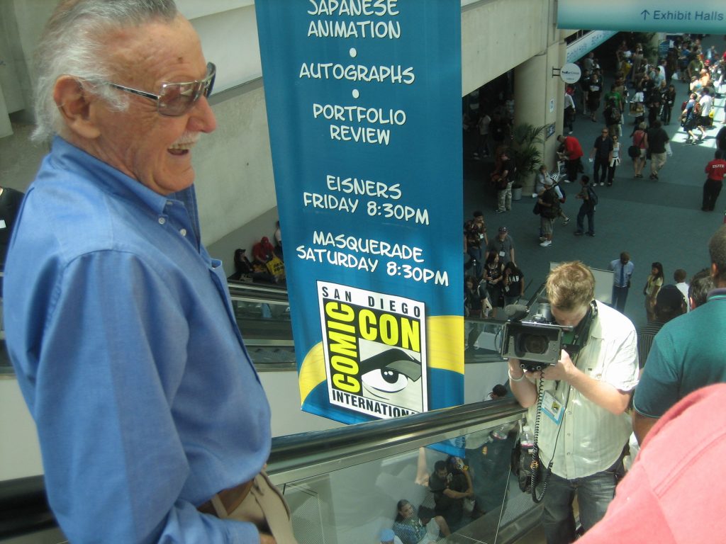 Stan Lee smiling while riding down an escalator at San Diego Comic Con with something filming him from below