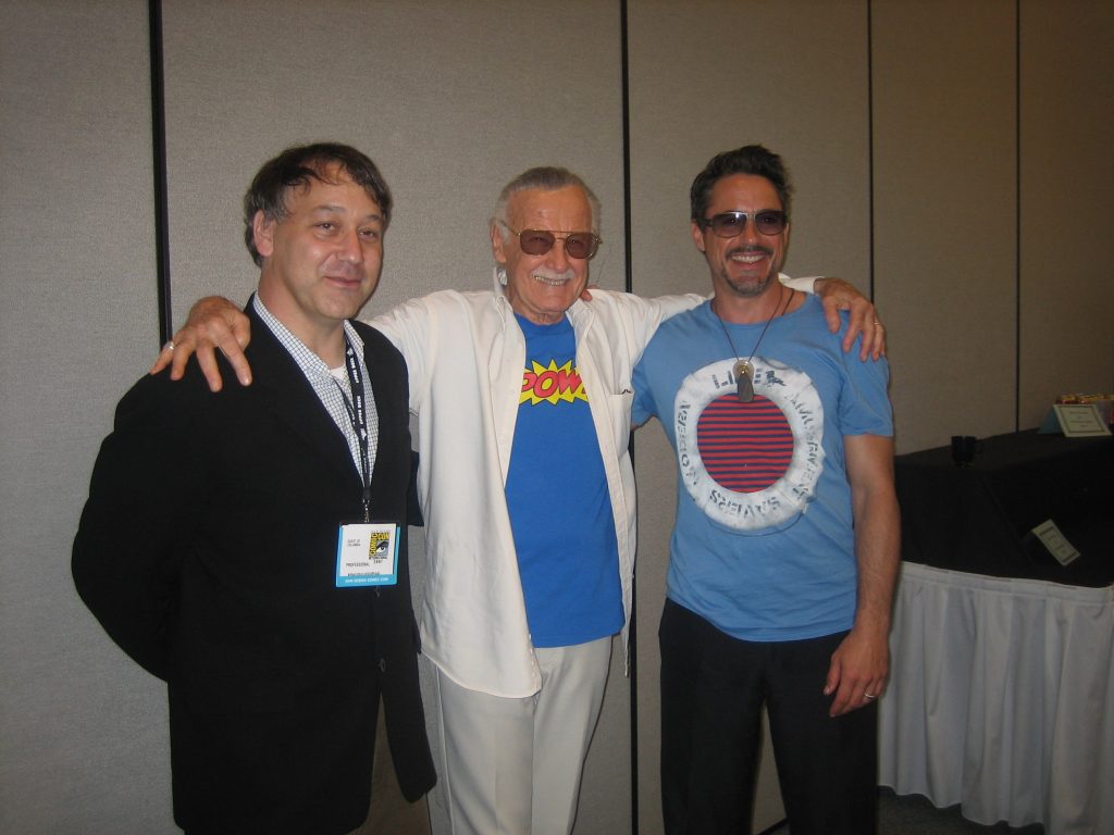 Stan Lee standing in the middle with his arms around the shoulders of Sam Raimi and Robert Downey Jr. at San Diego Comic Con 