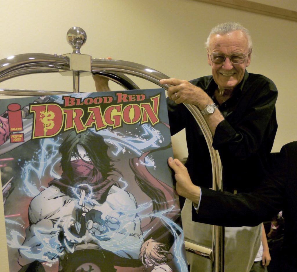 Stan Lee poses on a luggage cart with a poster for Blood Red Dragon at San Diego Comic Con 