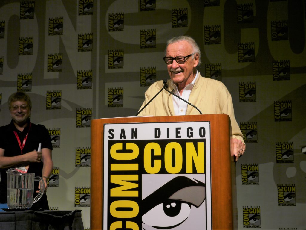 Stan Lee standing at a lectern and smiling during a panel at San Diego Comic Con 