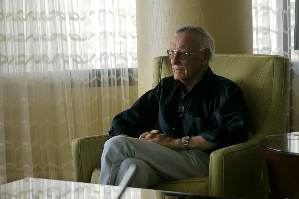 Stan Lee sits in a beige chair in a hotel lobby, looking off into the distance